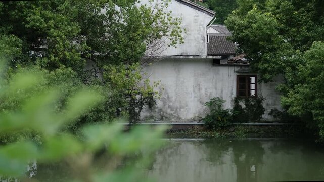 Weathered Jiangnan whitewashed house beside a river framed by lush foliage in rural Zhejiang China. Tranquil countryside waterway. Concept of cultural heritage, peaceful country life and rural tourism