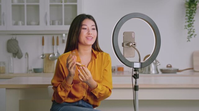 Smiling Asian Woman Holding Skincare Product, Sitting In Modern Kitchen With Ring Light. Happy Young Female Demonstrating Beauty Routine With Cosmetical Item, Emphasizing Self-care Concept.