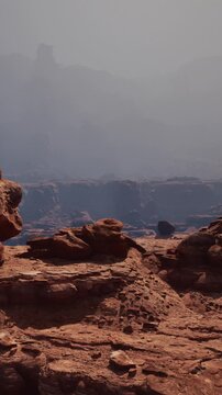 A group of large rocks stands on the ground in a desert area. In the background, cliffs and distant hills are visible under a bright sky. Shadows form on the ground.