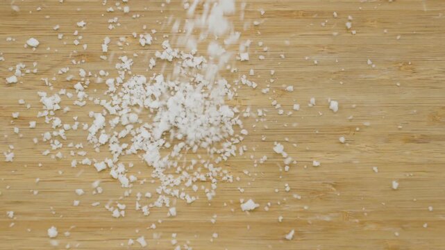 A close-up of white salt crystals scattered and spread across a wooden table surface with visible texture captured in slow motion.
