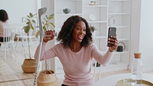 Smiling Black Woman Using Crutches While Taking Selfie With Smartphone At Home, Showcasing Positivity And Confidence, Sitting In Bright Modern Interior. Wellness And Healthcare Concept.