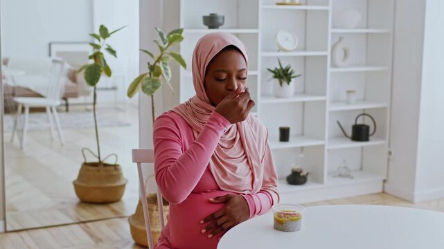 Focused Pregnant Black Woman Sitting At Kitchen Table, Enjoying Healthy Snack From Container, Nurturing Baby Bump In Bright Home Environment, Promoting Healthy Eating, Weight Loss Concept.