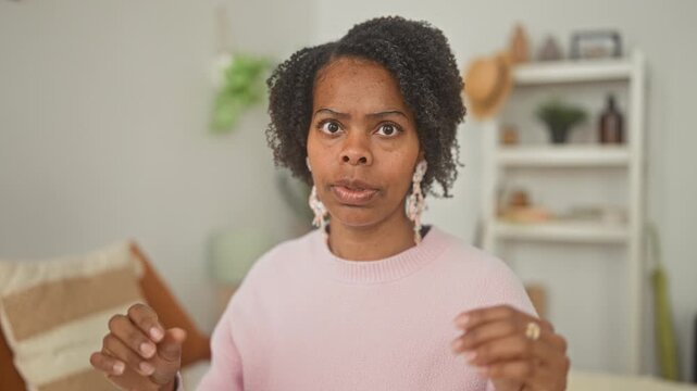 Woman gesturing with hands and expressive face on bed in studio, wearing pink sweater and statement earrings amid shelf and pillows; concern reflection.