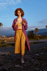 Fototapeta premium Vivid woman outdoors in colorful fashion, striped top, purple coat and mustard skirt, relaxed pose, sunlit scene, natural landscape, lifestyle portrait with bold styling