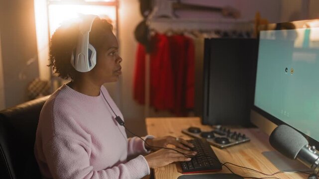 Young african american woman wearing white headphones typing on keyboard at wooden desk with monitor and audio mixer in studio; focus remote work.