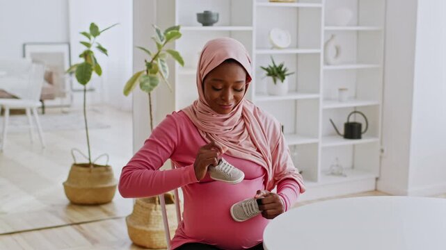 Smiling Black Woman In Pink Top Sitting At Home With Pregnant Belly, Excitedly Holding Baby Shoes, In Cozy Living Room, Preparing For Arrival Of Newborn. Maternity Concept.