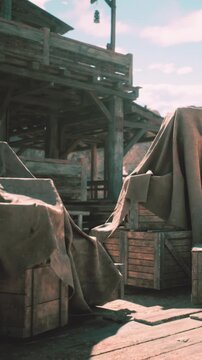 Wooden crates sit on a dock. They are covered with cloth and surrounded by rustic wooden structures. The sun shines on the scene, creating a bright atmosphere.