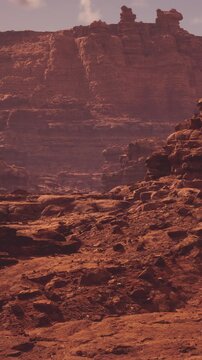 A desert scene shows a rocky terrain under a setting sun. The colors are warm as shadows form across the rocks. The landscape is vast and dry with no signs of water or vegetation.