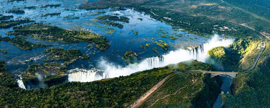 Aerial view of Victoria Falls, a massive waterfall on the Zambezi River, featuring lush vegetation and the Victoria Falls Bridge in Victoria Falls, Matabeleland North Province, Zimbabwe.