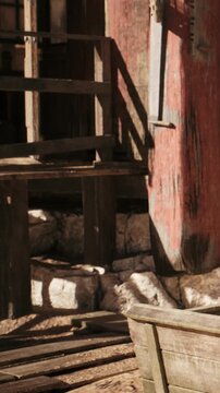 A wooden structure stands next to a stone ground. Red posts support the building. Shadows fall on the ground. Sunlight brightens the scene during the day.