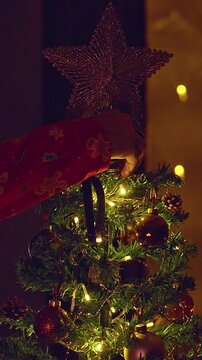 Vertical video. Person Placing a Glittering Star on Top of a Decorated Christmas Tree With Warm Fairy Lights and Fireplace Reflection in the Window, Cozy Holiday Tradition