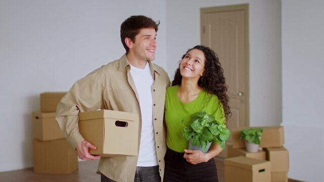 Beaming Young Couple, Smiling And Excited, Unpacking Boxes And Decorating Their New Home Together In A Cozy Living Room, Filled With Plants And Moving Boxes. Home And Lifestyle Concept.
