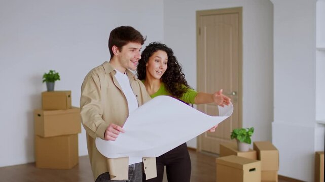 Smiling Young Couple, One Male And One Female, Holding Architectural Floor Plan And Discussing Design Ideas In Their New Modern Apartment, Excited About Home Renovation Project.