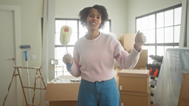 Young woman holding a paint roller and smiling while unpacking cardboard boxes in an apartment, wearing pink sweater and jeans, preparing a living room for renovation; moving day joy.
