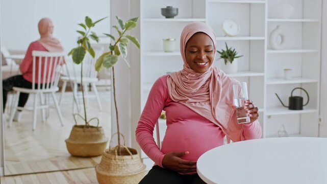 Happy Pregnant Woman With Pink Hijab Sitting At Home, Gently Cradling Her Baby Bump And Enjoying A Glass Of Water, Feeling Content And Relaxed In A Bright, Cozy Living Room. Expecting Mother
