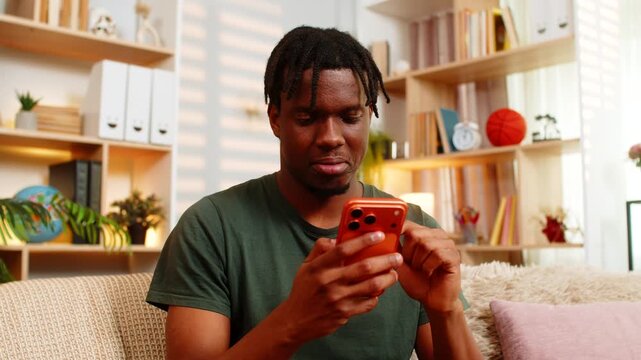African American young man on home sofa reading message on smartphone with relaxed smile. Black guy on home sofa texting in living room feeling happy calm focused during mobile chat in indoor scene.