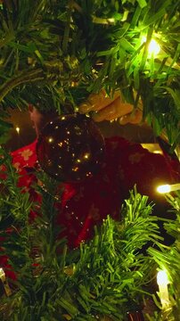 Vertical video. Creative View From Inside a Decorated Christmas Tree Framing a Young Woman Decorating Holiday Ornaments With Warm Fairy Lights and Cozy Fireplace Reflections