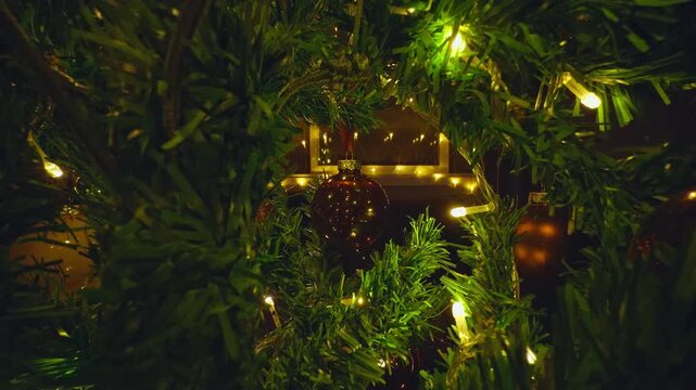 Christmas Tree Branches and Glowing Baubles Framing Warm Window Lights and Cozy Holiday Interior in a Creative Point of View Shot