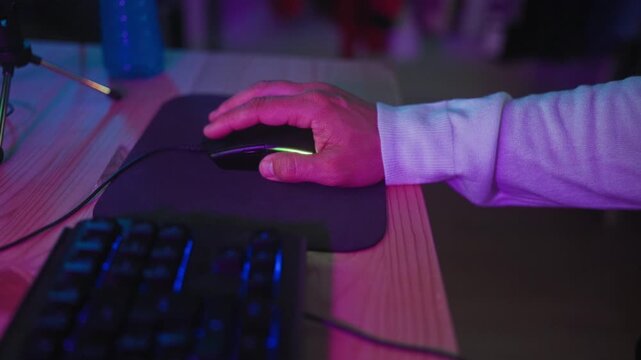 Man hand holds gaming mouse on mousepad and clicks near keyboard at studio desk under purple led lighting, visible cable and microphone stand; concentration gaming.