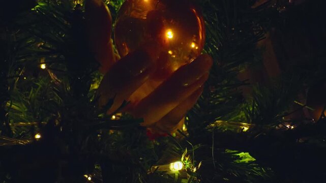 Close Up From Inside a Christmas Tree as Female Hands Hang a Golden Holiday Bauble Among Warm Fairy Lights and Evergreen Branches