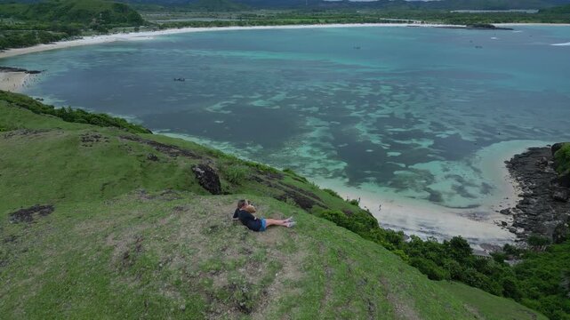 Caucasian Backpacker Seated Above Coral Reef, Gazes At Shallow Turquoise Waters And Sandy Inlet, Panoramic Coastal Vista With Calm Reflective Atmosphere And Midday Light, Bukit Merese, Lombok