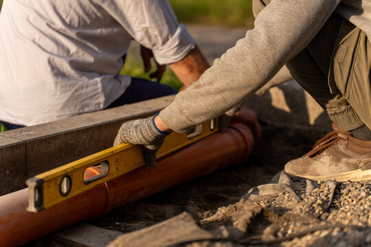 Worker using a yellow spirit level to check the slope of an orange plastic sewage pipe in a ground trench on an outdoor construction site.