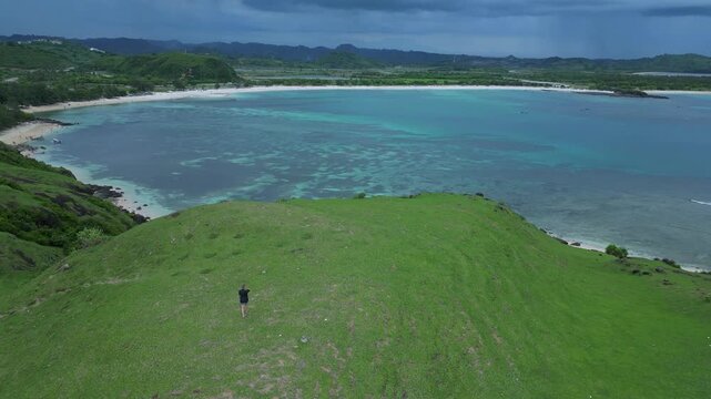 Woman Surveying Wide Turquoise Bay From Hilltop With Expansive Lagoon And Distant Shoreline Solitary Figure On Crest, Curving Paths In Grass, Calm Sea Tones, Cinematic Aerial Vista, Lombok