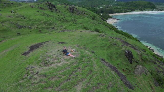 Traveler and her dog View Coast. Hiker Pauses To See Landscape. Explorer Stops To Admire Vibrant Shoreline And Sea. Wanderer Takes Break Atop Hill To Enjoy Lively Bay And Rocky Coast, Lombok