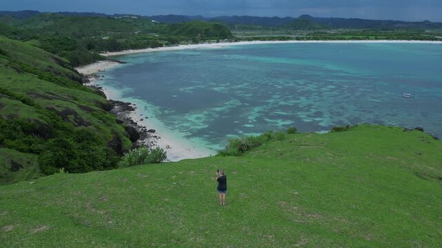Woman Surveying Wide Turquoise Bay From Hilltop With Expansive Lagoon And Distant Shoreline Solitary Figure On Crest, Curving Paths In Grass, Calm Sea Tones, Cinematic Aerial Vista, Lombok