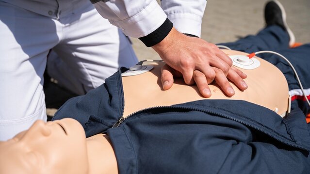 A medical professional performs simulated chest compressions on a CPR training mannequin outdoors.