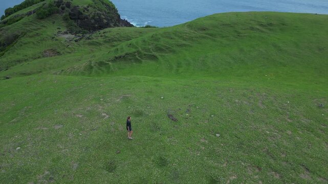Woman Descending Coastal Cliff Path Toward Beach And Rocky Shoreline Narrow Trail, Surf Lines, Textured Grass Slopes, Overcast Light, Aerial Framing Conveys Journey And Scale, Bukit Merese, Lombok