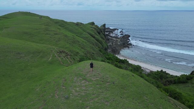 Woman Descending Coastal Cliff Path Toward Beach And Rocky Shoreline Narrow Trail, Surf Lines, Textured Grass Slopes, Overcast Light, Aerial Framing Conveys Journey And Scale, Bukit Merese, Lombok