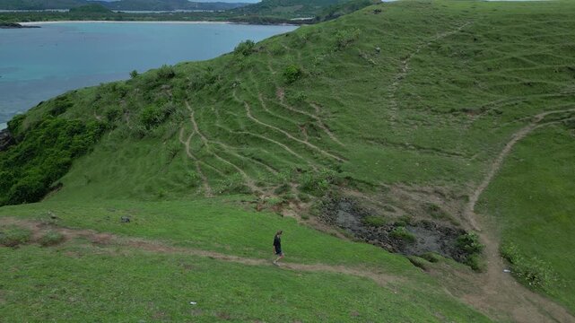 Woman Walking Cliffside Path Toward Headland, Rocky Promontory And Open Sea Frame Steady Forward Stride, Expansive Coastal Vista, Travel And Adventure Tone With Rugged Green, Lombok