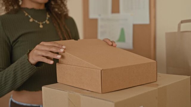 Woman holding cardboard parcel with hand on lid while placing it on stacked shipping boxes in studio; small business confidence.