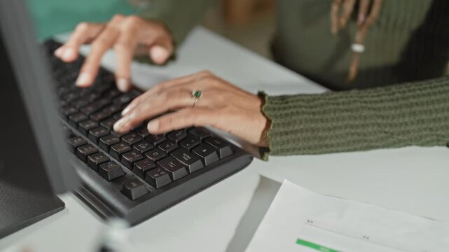 Woman typing on keyboard with visible hands and ring, wearing green sweater at office desk in building; concentration.