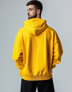 Person standing with back to camera wearing bright yellow hoodie and black pants, hands in pockets, calm introspective mood against a neutral gray studio background