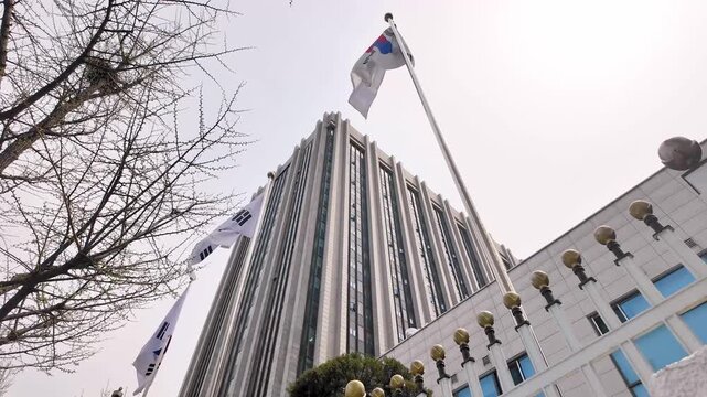 A low angle view looking up at a tall government building with national flags and bare tree branches against a cloudy sky.