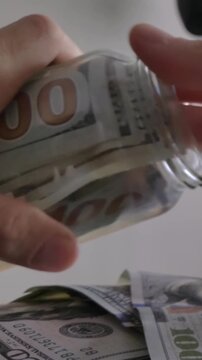 Man Stashing Hundred Dollar Banknotes In Glass Piggy Bank For Savings - 4K Vertical