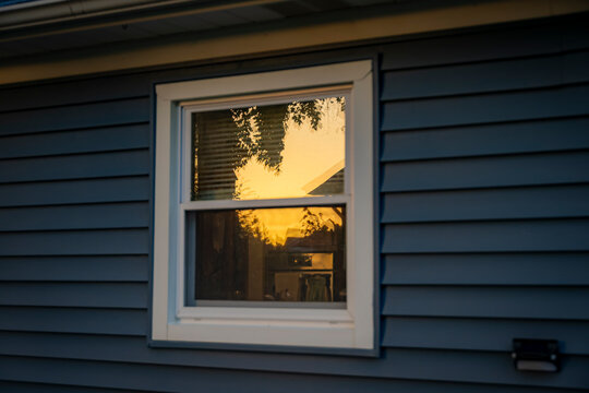 Golden sunset sky and tree silhouettes reflected in a glass window pane of a house with blue vinyl siding close up view