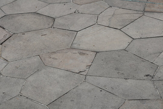 A weathered outdoor pavement made of large concrete pavers. The flagstones are cut into irregular, geometric polygonal shapes fitted tightly together with thin grout lines.