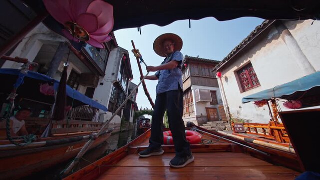 A gondolier expertly maneuvers a traditional boat through the narrow canals of Zhouzhuang. Sunlight highlights the historic architecture. Ideal for cultural documentaries or travel features.