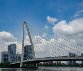 Obraz premium Modern Cable-Stayed Bridge Over a River With City Skyline and Blue Cloudy Sky