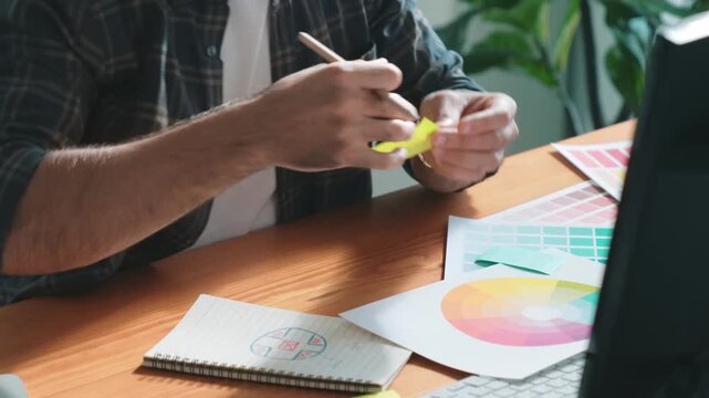 Professional designer writing and designing logo graphic at meeting. Closeup of business man working on colored palettes and writing idea on sticky notes on table with equipment and paper. Symposium.