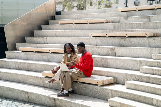 Nerdy african american man student helping black female friend with studies, explaining work process on laptop, black male and woman graduates sitting outdoors at university using computer and talking