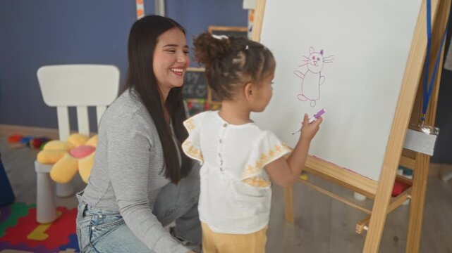 Woman helps little girl draw on easel, hand on back while the child sketches a cat on a whiteboard in studio for art practice; learning bonding joy.