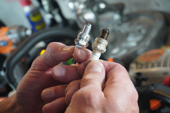 A close-up shot of a person's hands holding a clean new spark plug and a dirty fouled spark plug side-by-side in a workshop, demonstrating engine maintenance and auto repair.