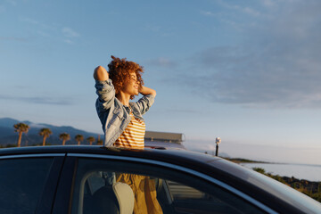 A young woman leans out of a car window, hair blowing in the wind against a coastal sunset, conveying freedom and carefree summer energy. © SHOTPRIME STUDIO