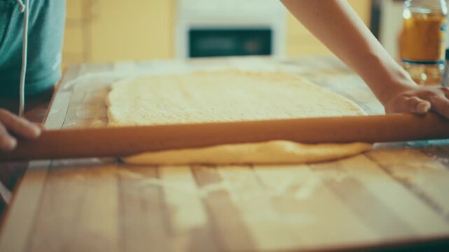 Close-up of baker hands using a wooden rolling pin to roll out and flatten fresh yeast dough. Preparing homemade sweet pastry or cinnamon rolls on a floured table, 4k.