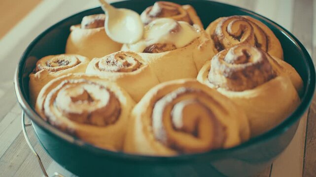 Close-up of chef hand spreading white cream glaze with a spoon onto freshly baked hot cinnamon rolls in a baking pan. Preparing sweet homemade pastry dessert on a wooden background, 4k.