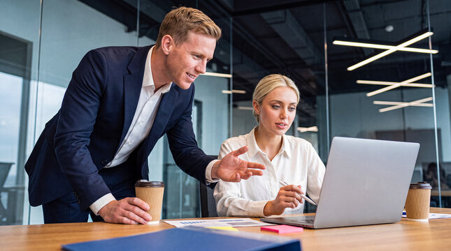 Professional male colleague mentoring or assisting a female coworker with a laptop in a sleek modern office environment.
